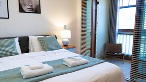 Ein ordentlich bezogenes Bett mit Kissen, einer Nachttischlampe und Handtüchern. Im Zimmer befindet sich ein Sessel und ein Fenster mit Jalousien.