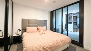 Une chambre moderne avec un grand lit, une table de chevet, une lampe et une porte-fenêtre coulissante donnant sur un balcon extérieur.