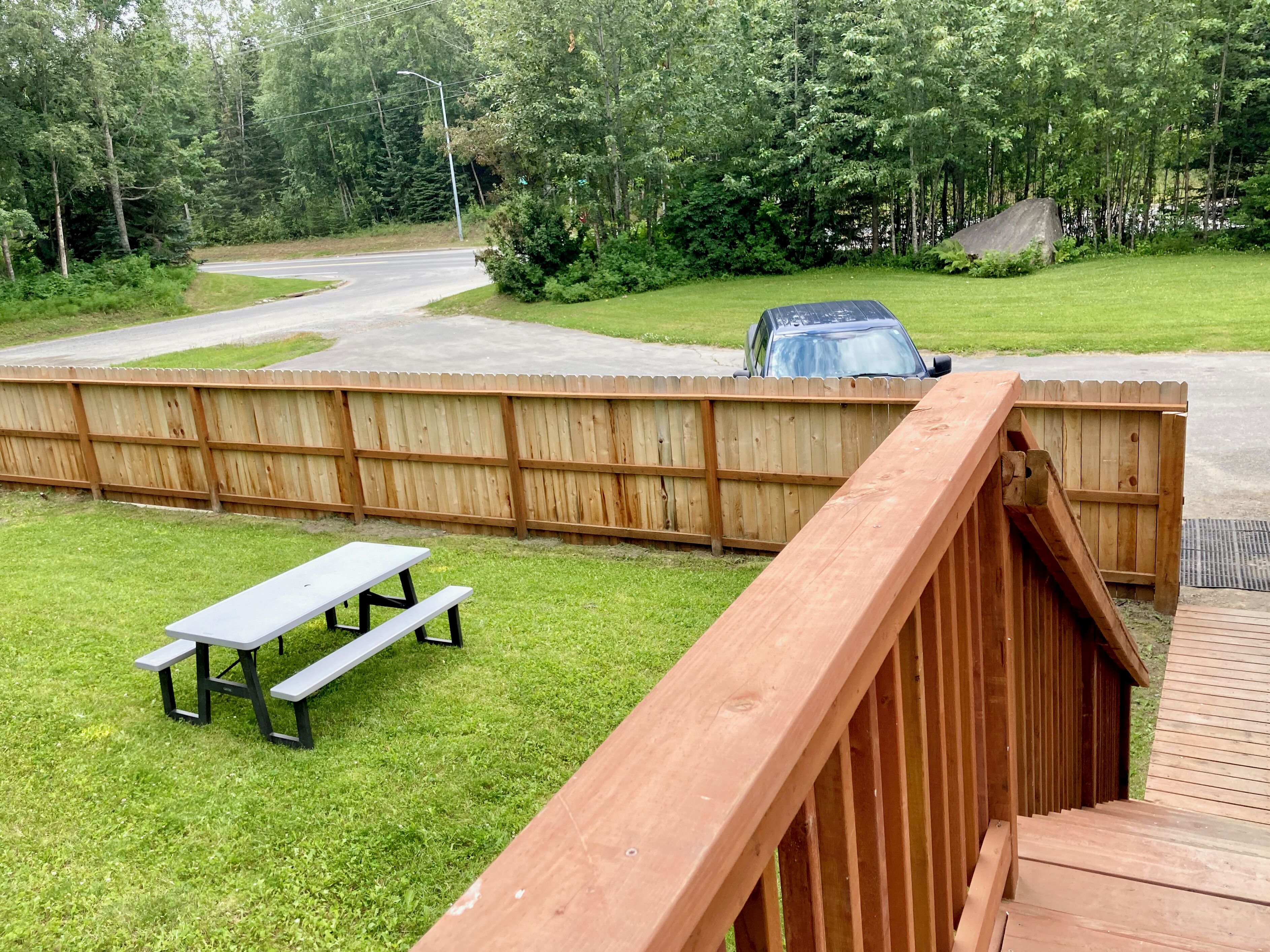 A wooden deck with a picnic table, a fenced yard, and a road in the background.