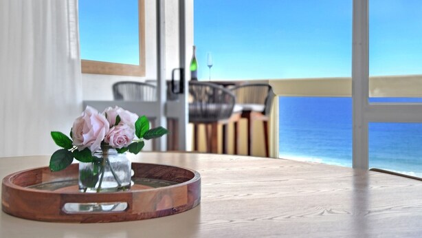 A table with a wooden tray holding a vase of pink roses, a dining area with chairs, and a view of the ocean through large windows. - Breathtaking Ocean view 3Bedrooms Unit next Beach (Surfers Paradise)