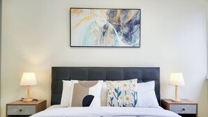 Una cama bien hecha con una colcha blanca y varios almohadones, flanqueada por dos mesitas de noche con lámparas, en una habitación con un diseño sencillo y limpio.