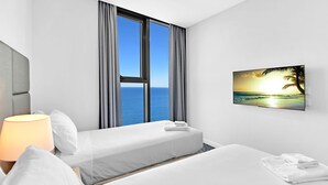 Ein Hotelzimmer mit zwei Betten, einem großen Fenster mit Meerblick, einem Sonnenuntergangsgemälde und einer Lampe auf der linken Seite.