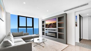 Ein modernes Wohnzimmer mit großem Fenster zum Meer, einem grauen Sofa, einem Glastisch und einer eingebauten Regalanlage mit Fernseher.
