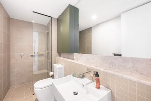 Un baño moderno con cabina de ducha de vidrio, inodoro blanco, lavabo con dispensador de jabón y espejo sobre el lavabo.