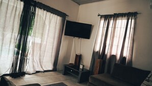 A living room with a flat-screen TV mounted on the wall, a sofa, a small table with a basket, and a window with sheer curtains.