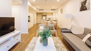 Un salon moderne avec un téléviseur à écran plat, un canapé gris, une table basse agrémentée d’une plante, et un coin repas à l’arrière-plan.