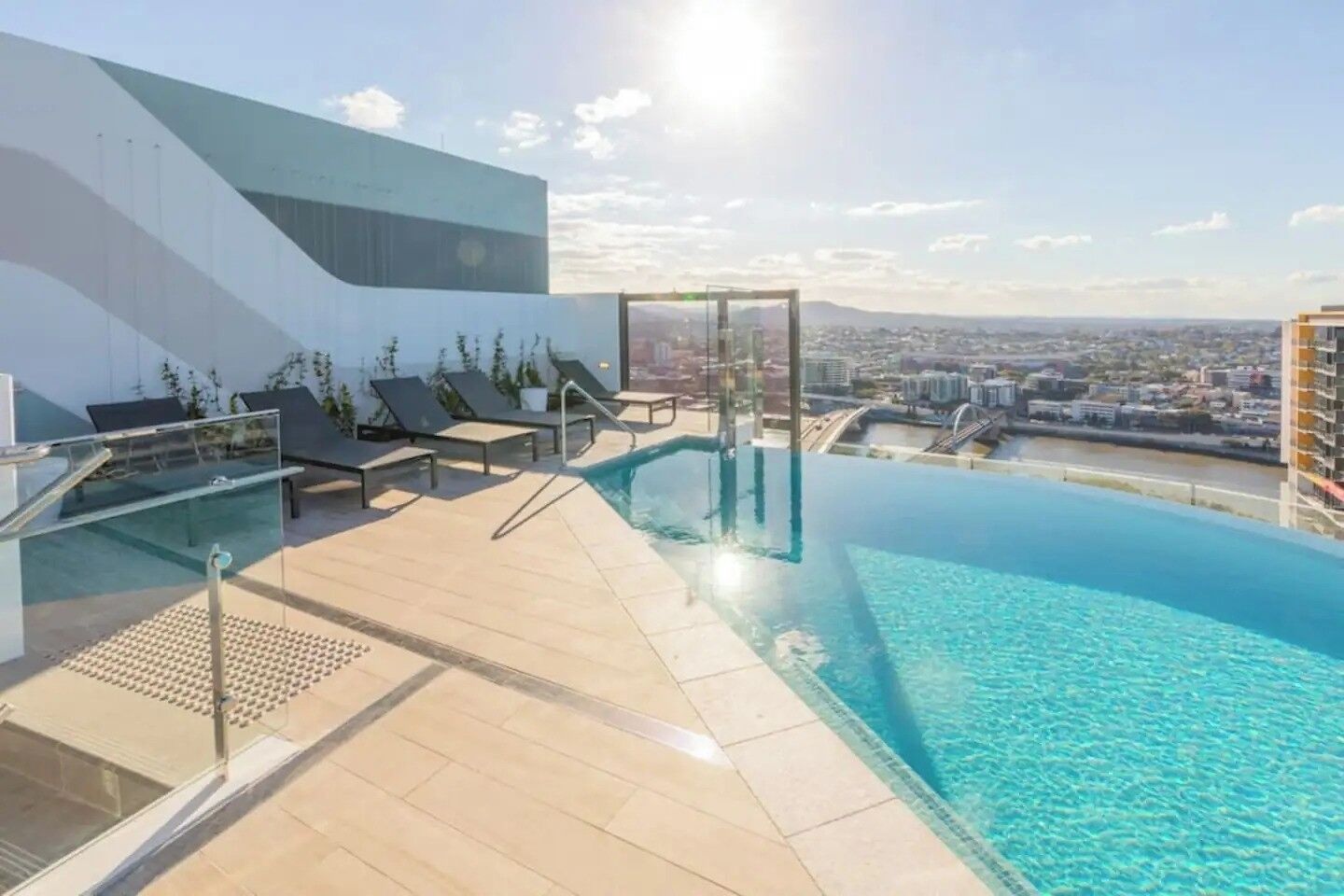A rooftop infinity pool with lounge chairs and a glass balustrade, overlooking a cityscape.