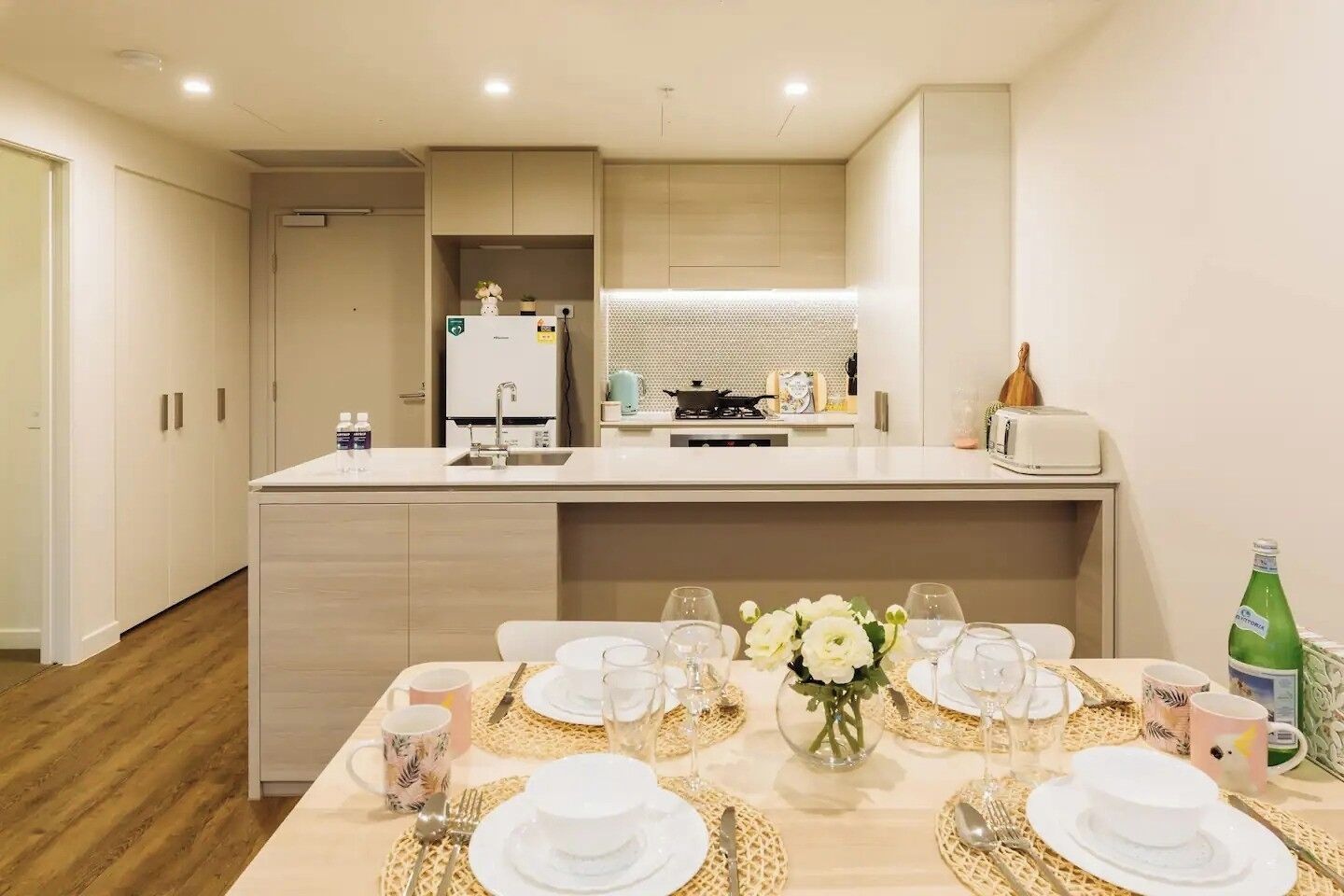 A modern kitchen with a central island, a dining area set for four, and a small refrigerator.