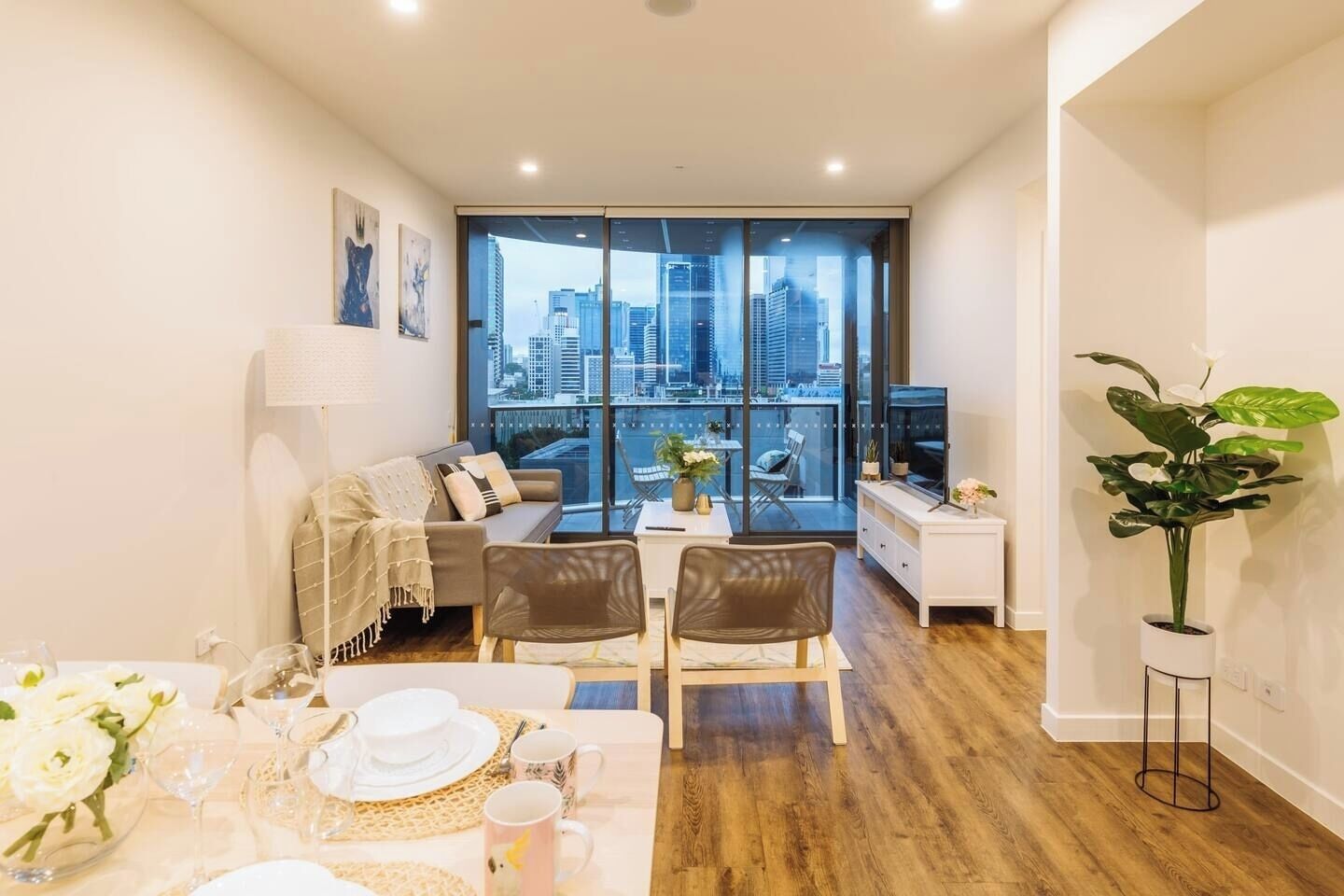 A modern living room with a dining area, large windows offering a city view, a sofa, a TV, and a potted plant.