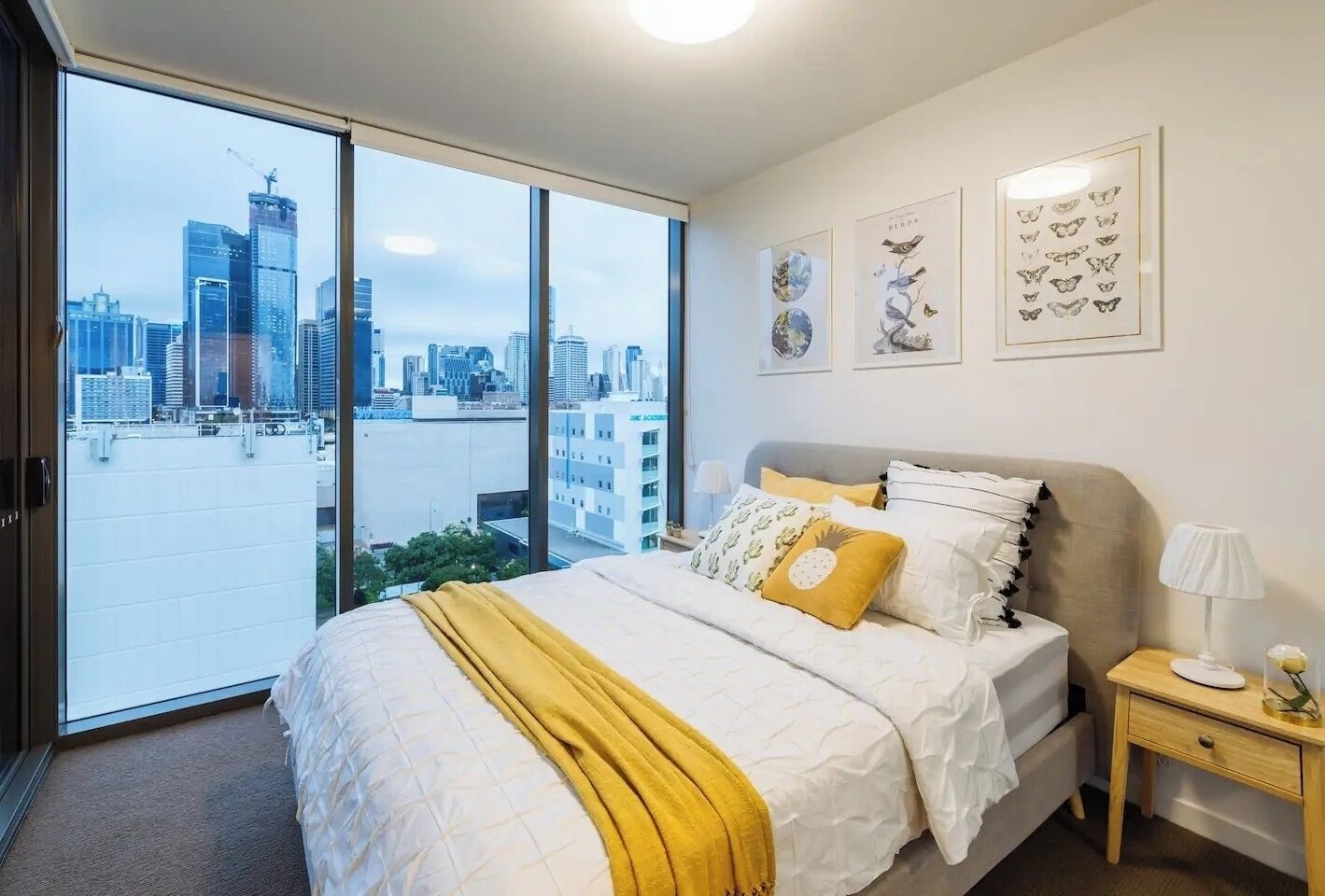 A bedroom with a large bed, a nightstand, a lamp, and a view of a cityscape through large windows.