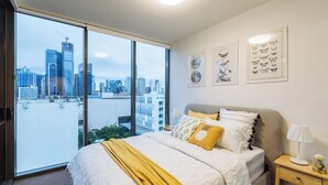 Une chambre à coucher avec un grand lit, une table de chevet, une lampe et une vue sur le paysage urbain grâce à de grandes fenêtres.
