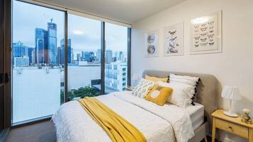 A bedroom with a large bed, a nightstand, a lamp, and a view of a cityscape through large windows.