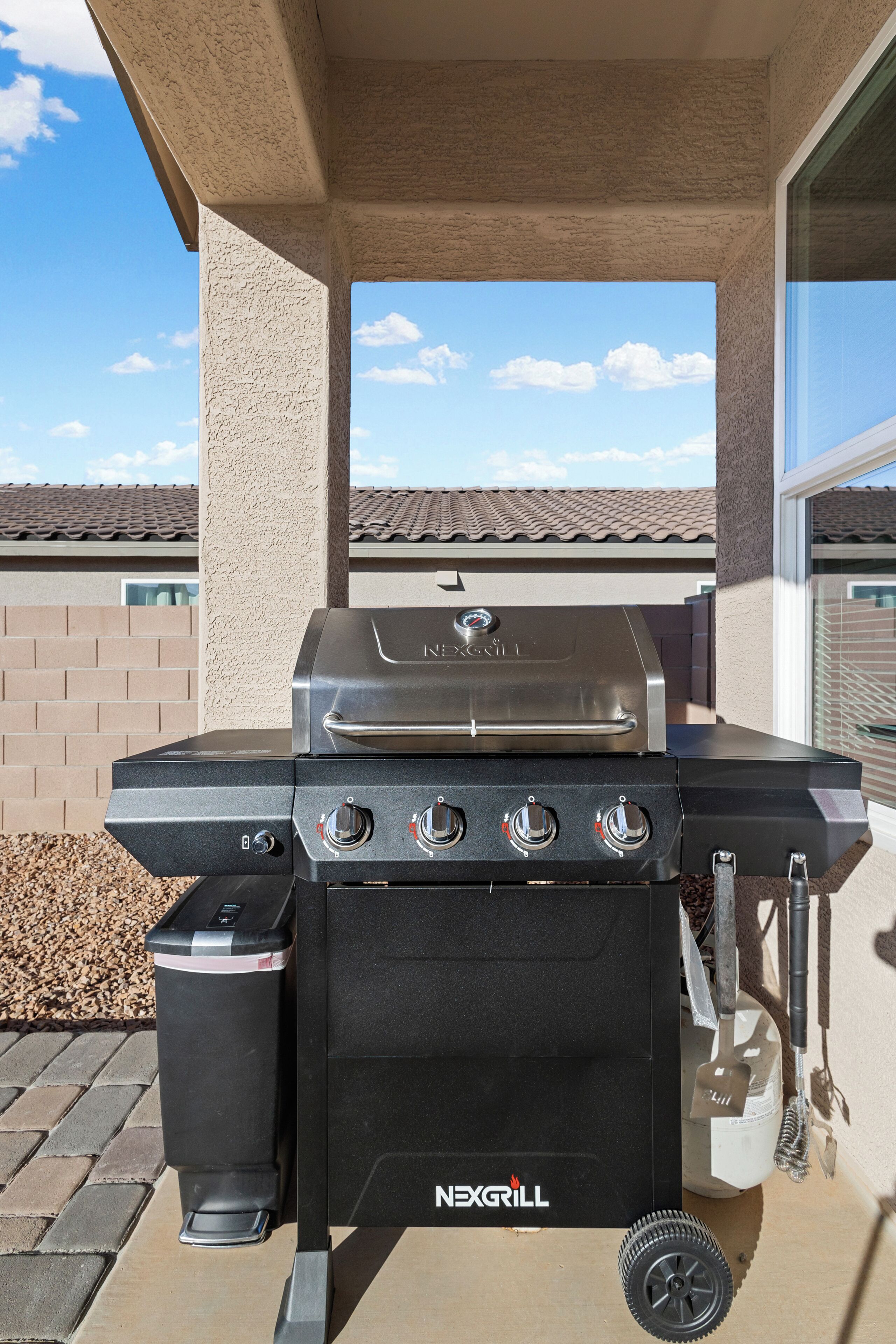 Een NEXGRILL barbecue met vier branders, een bijkomende brander en een waterreservoir, staand op een terras met een helder uitzicht op de lucht en een betegeld dak op de achtergrond.