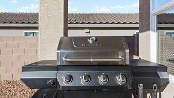 Een NEXGRILL barbecue met vier branders, een bijkomende brander en een waterreservoir, staand op een terras met een helder uitzicht op de lucht en een betegeld dak op de achtergrond.
