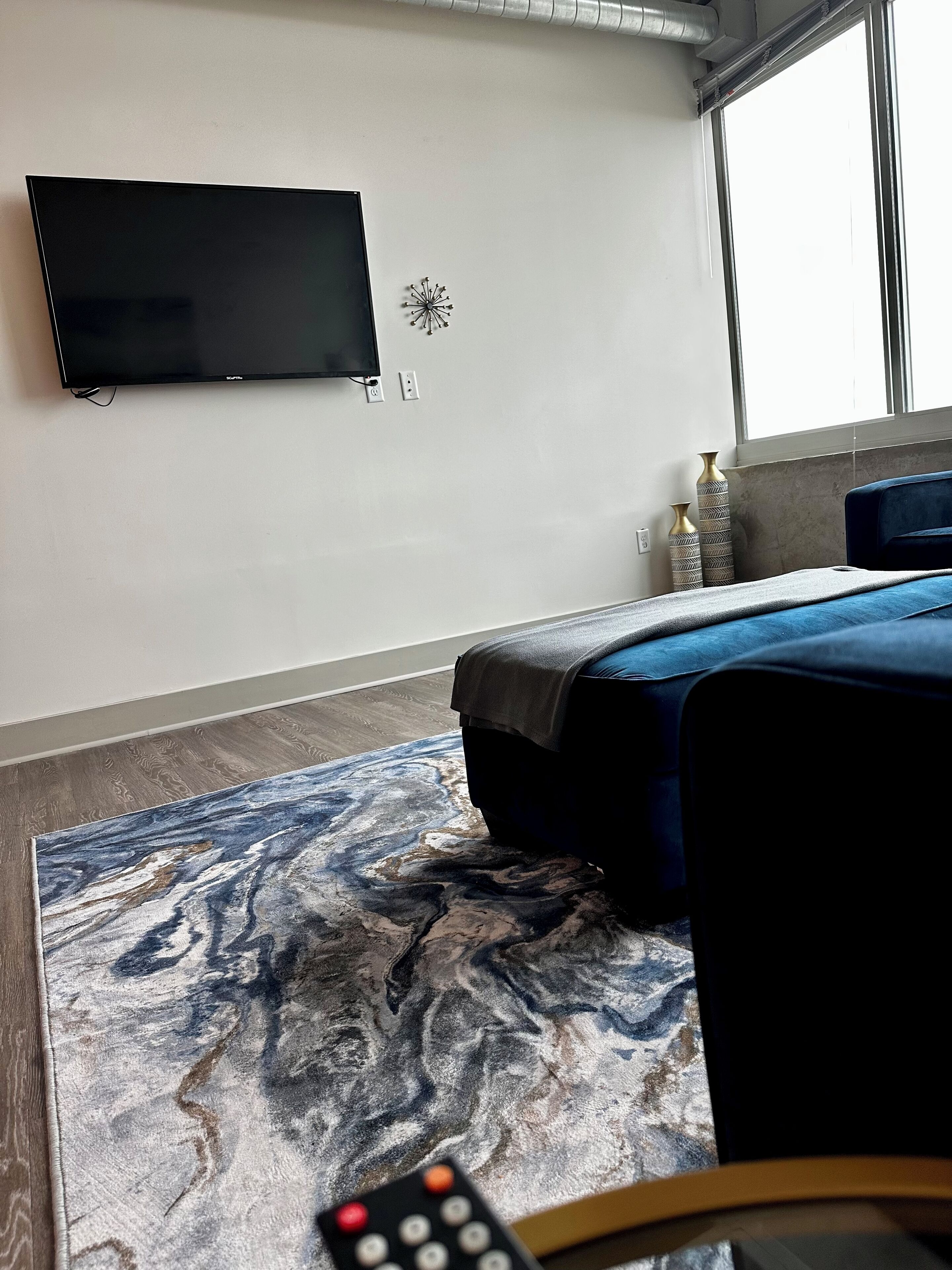 A modern hotel room with a flat-screen TV mounted on the wall, a bed with a blue headboard, a marble-patterned rug, and a remote control on the floor.