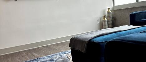 A modern hotel room with a flat-screen TV mounted on the wall, a bed with a blue headboard, a marble-patterned rug, and a remote control on the floor.