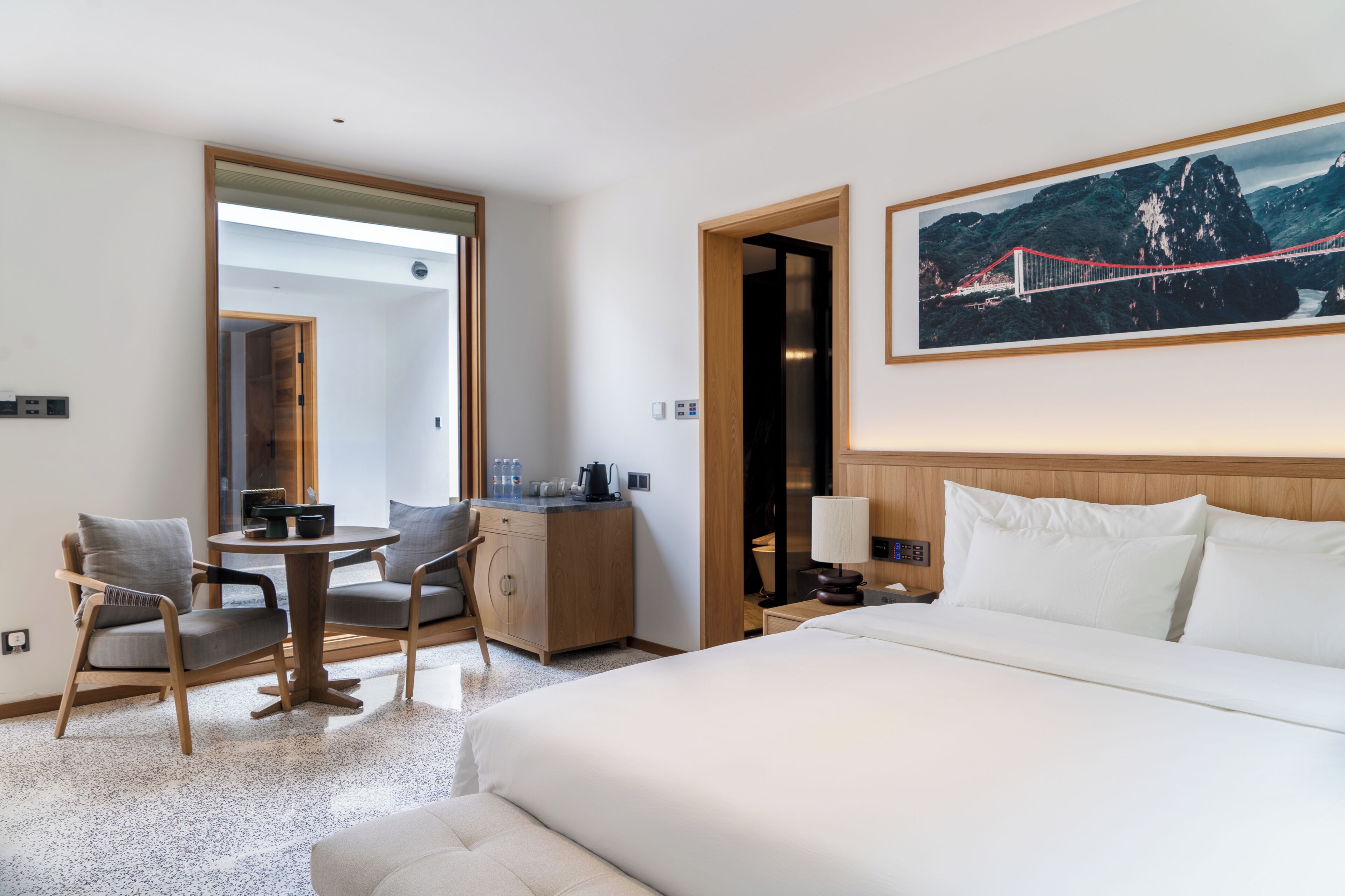 A hotel room with a bed, a desk with a chair, a small table, and a painting of a bridge.