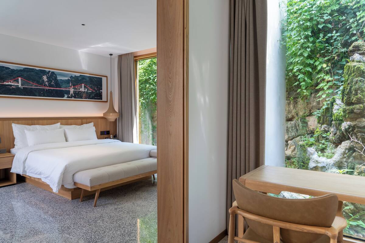 Ein modernes Hotelzimmer mit einem großen Bett, einem Holztisch, Blick ins Grüne durch ein Fenster und einem gerahmten Bild an der Wand.