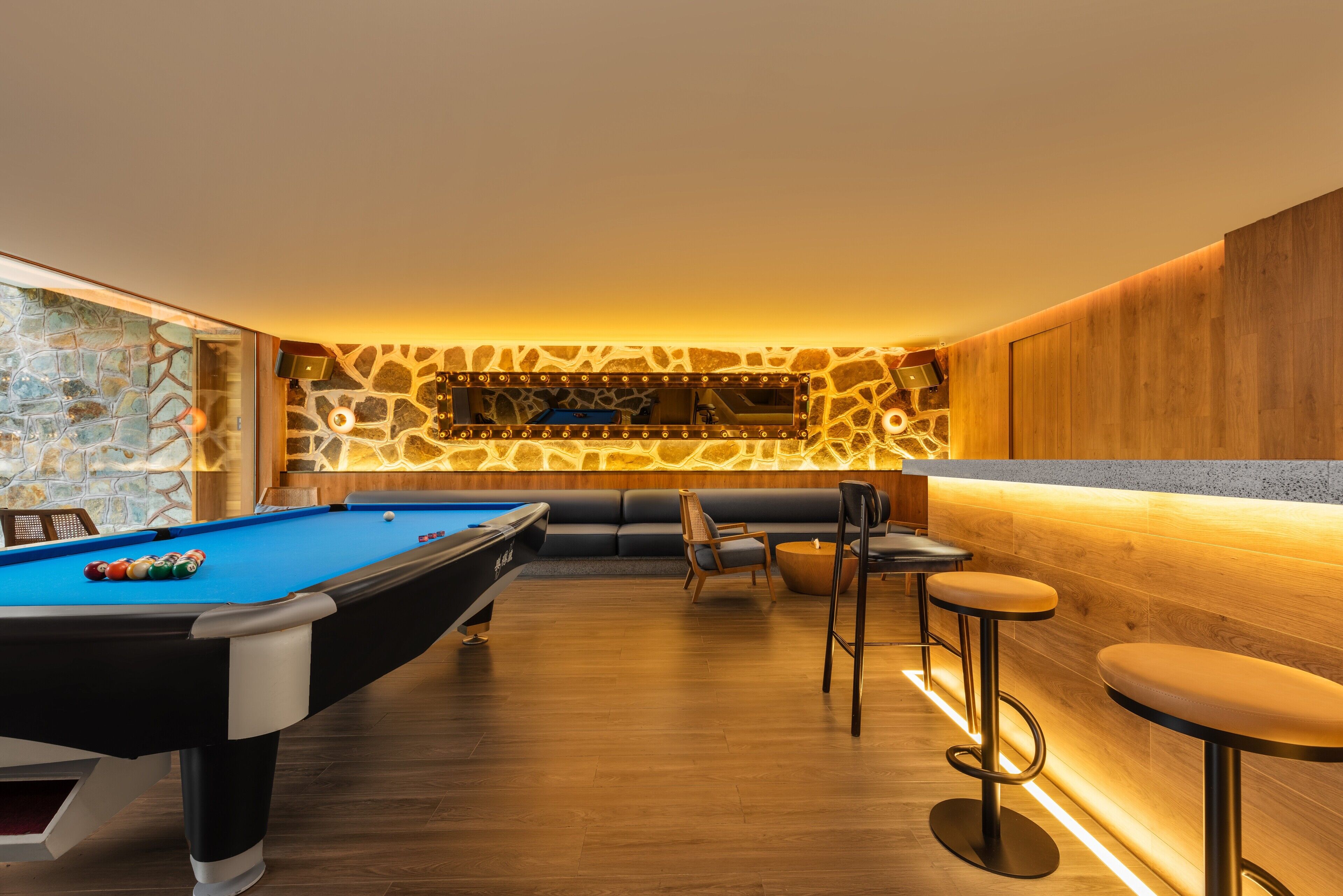 Une table de billard recouverte d’un feutre bleu, un bar en bois avec des tabourets et un mur décoré d’un motif imitant la pierre.