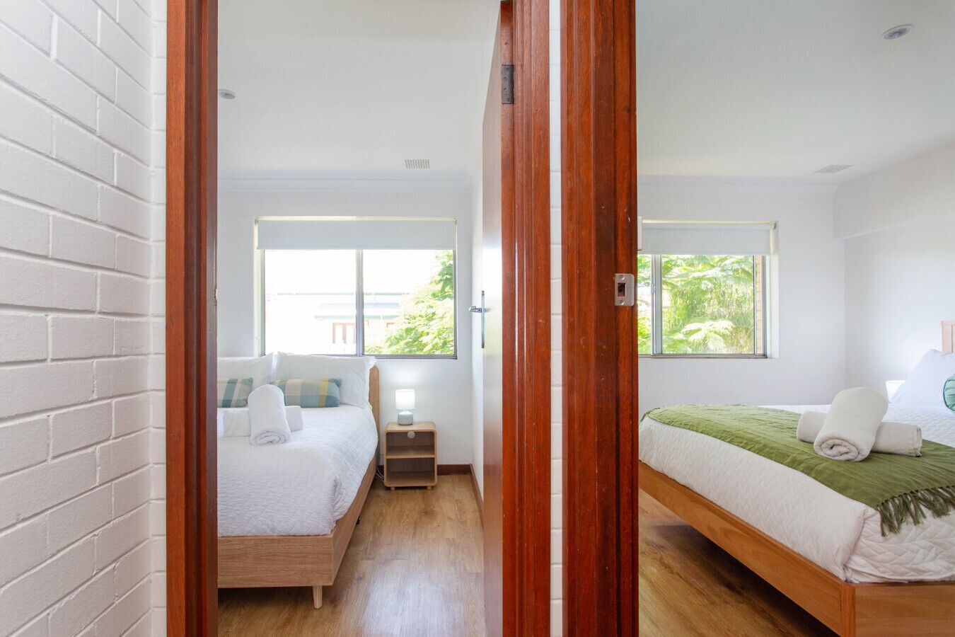 A hotel room with two beds, wooden flooring, a brick wall, and large windows with a view of greenery.