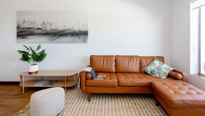 A modern living room with a brown leather sofa, a white brick wall, a large abstract painting, a wooden coffee table, and a potted plant. - Modern Fremantle- Sunset Over the Ocean (Fremantle)
