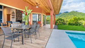 Un área de comedor exterior moderna con piscina, techo de madera y vista a un paisaje verde exuberante.