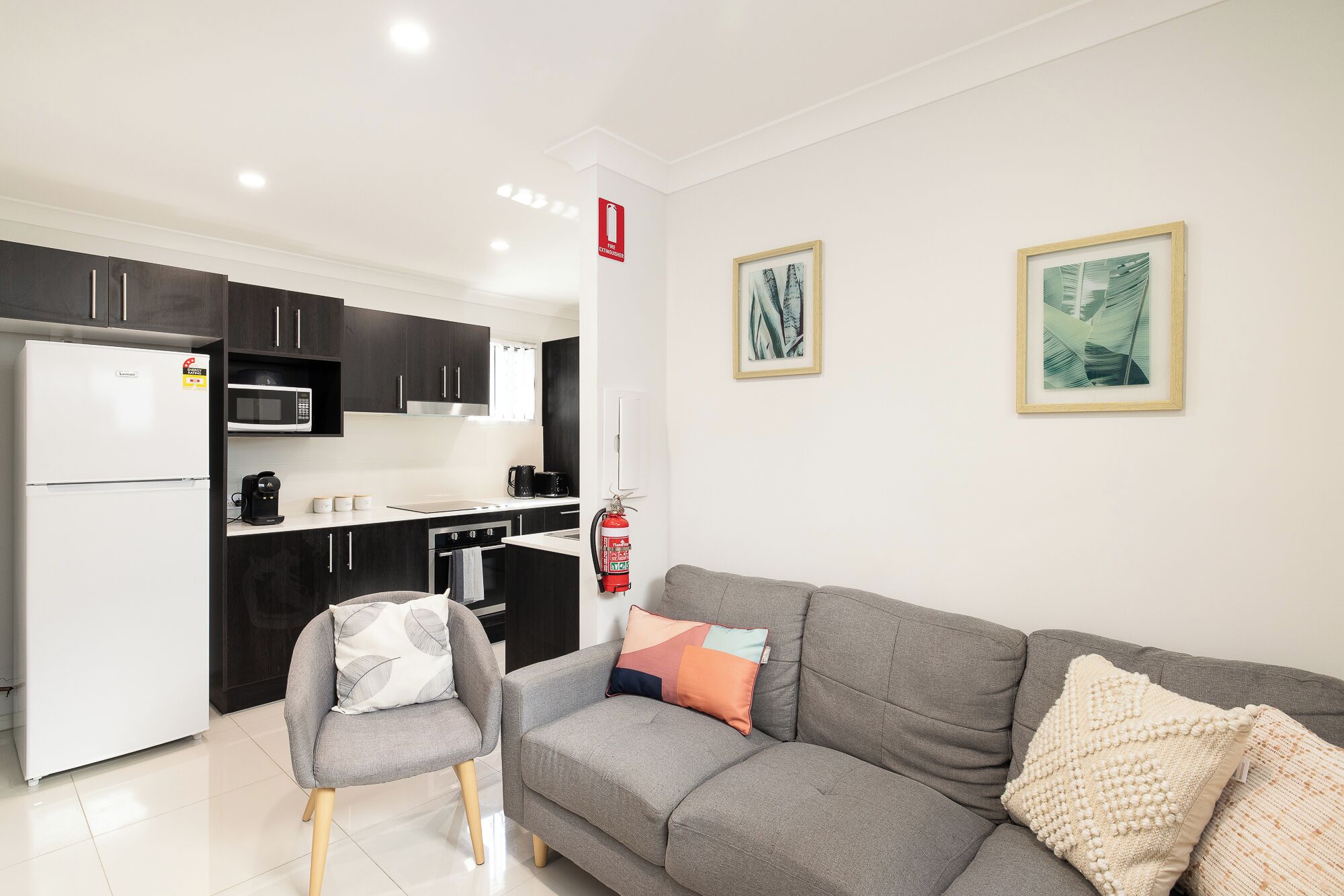 A modern kitchen with dark cabinets, a white refrigerator, and a microwave. There is a sofa with cushions and a fire extinguisher mounted on the wall.