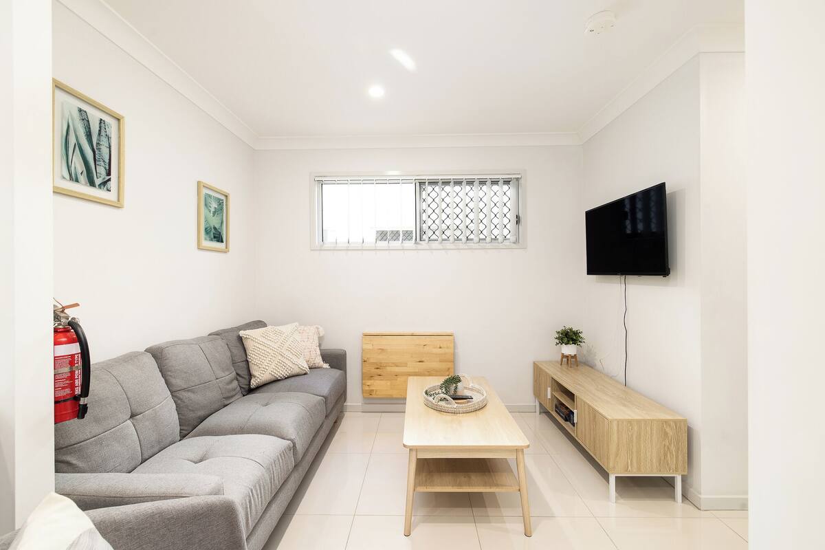 Un salon moderne avec un canapé gris, une table basse en bois, un téléviseur fixé au mur et une fenêtre ornée d’un motif en treillis.