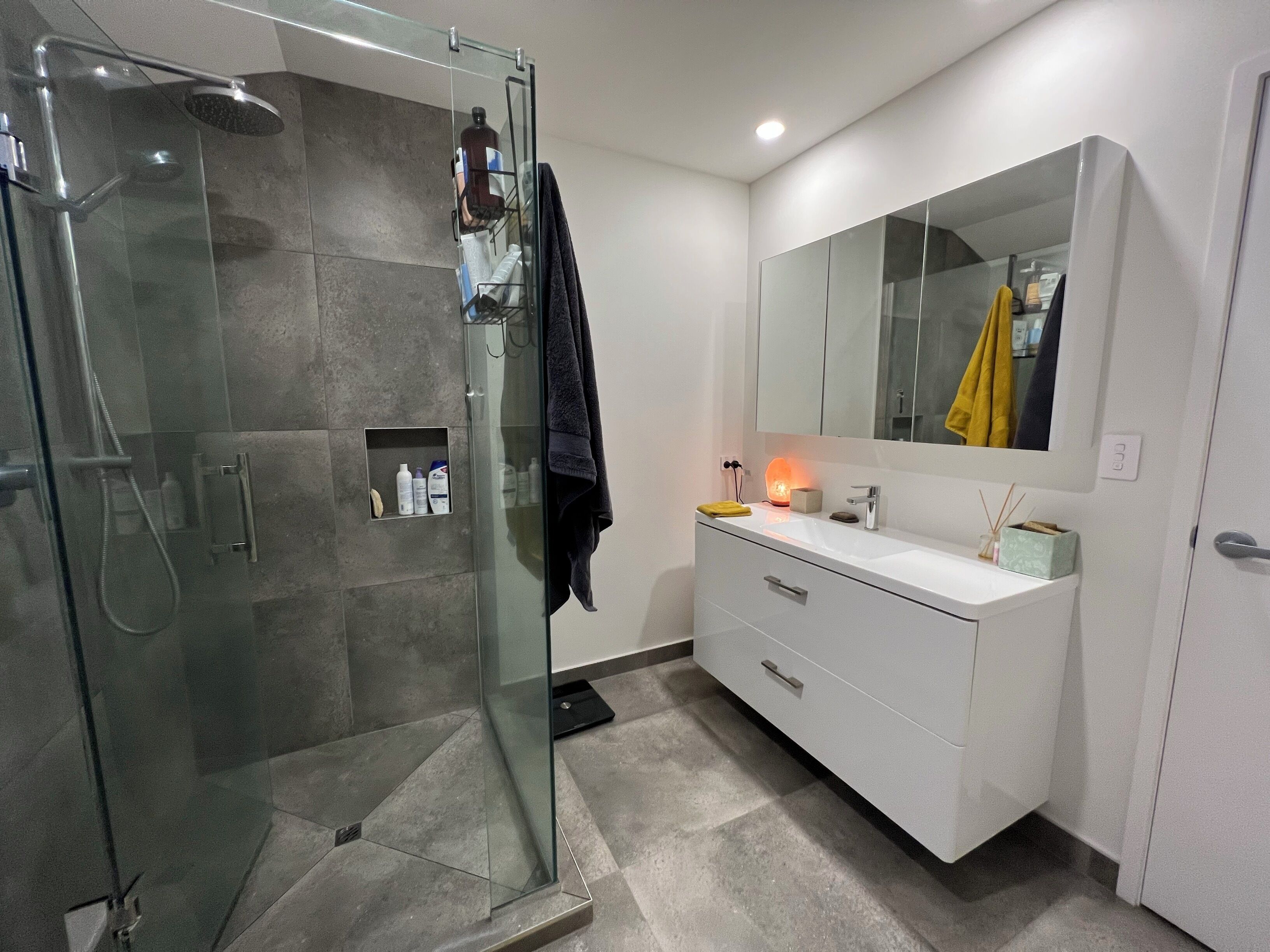 A modern bathroom with a glass-enclosed shower, a white vanity with a mirror, and a small shelf with toiletries.