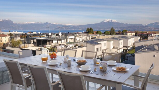 A dining table set with breakfast items and a scenic view of a coastal town with mountains in the background. - Perla Garda Lake Apartment With Pool (Peschiera del Garda)