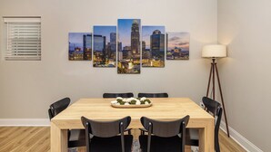 A dining area with a wooden table, black chairs, a cityscape artwork on the wall, a floor lamp, and a window with blinds.