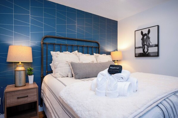 A hotel room with a blue tiled wall, a metal-framed bed, two bedside lamps, a framed horse picture, and a neatly made bed with towels and a robe.