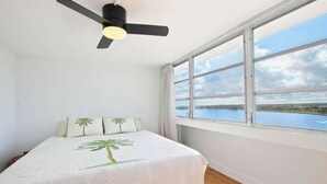 A bedroom with a bed, a ceiling fan, and a large window with a view of the sea.