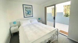 A bedroom with a bed, a nightstand, a framed picture on the wall, and a sliding glass door leading to a balcony. - Central location | Alfresco dine on huge terrace | Walk to cafes/shops/blowhole (Kiama)