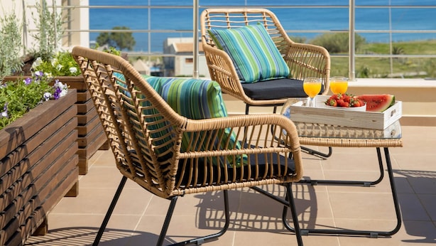 A balcony with wicker chairs, a small table with fruit and drinks, and a view of the sea.