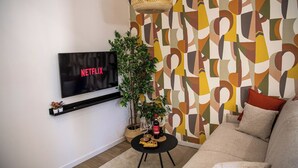 Una sala de estar con un televisor de pantalla plana en la pared, una pequeña mesa redonda de centro con una botella y vasos, una planta en maceta y un sofá con cojines.
