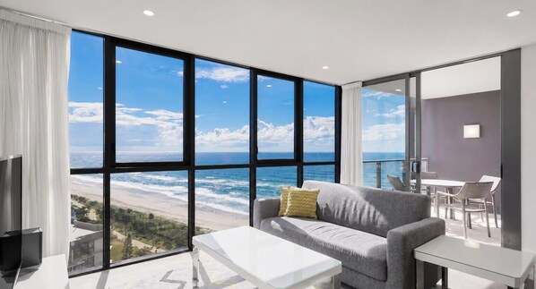 A modern room with a large window overlooking the ocean, a grey sofa, a white coffee table, and a dining area with white chairs.