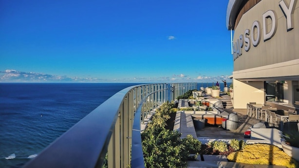 Un balcón con mobiliario exterior, vistas al mar y un edificio con el rótulo 'HYSODY'.