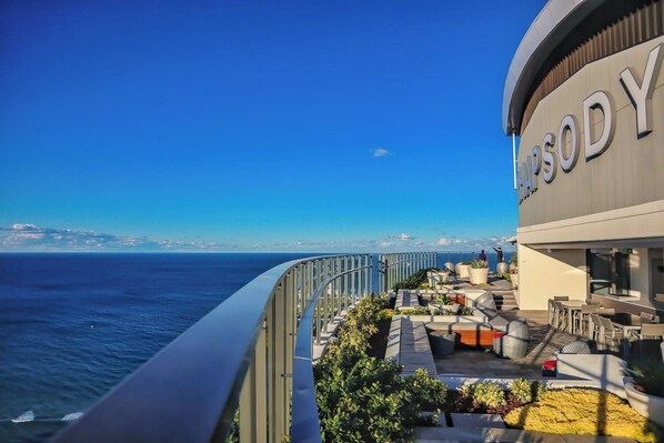 Un balcon avec des sièges d’extérieur, une vue sur la mer et un bâtiment portant l’enseigne « HYSODY ».