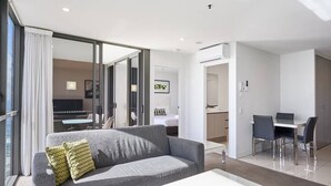 Una habitación de hotel moderna con un sofá gris, una mesa de centro blanca, un área de comedor con una mesa y sillas, y un dormitorio separado con una cama y un escritorio.