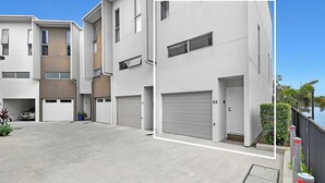 A modern multi-story residential building with a clean, minimalist design, featuring a spacious driveway and a view of the water. - Tri-Level 2BR Water-View Townhouse | Free Parking (Carrara)