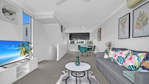 A modern living room with a flat-screen TV, a grey sofa with tropical cushions, a round coffee table, and a dining area with teal chairs. - Tri-Level 2BR Water-View Townhouse | Free Parking (Carrara)