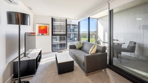 A modern living room with a sofa, coffee table, TV, and large windows offering a view of the cityscape. - Modern 1 Bedroom Apartment in Philip Canberra (Phillip)
