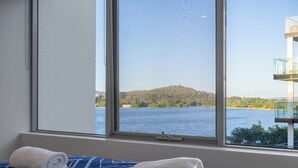 Een kamer met een bed, een raam met uitzicht op een meer en een heuvel, en een balkon met een leuning.