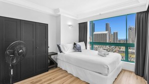 Ein modernes Hotelzimmer mit einem großen Bett, einem Standventilator und Blick auf Stadtgebäude durch ein großes Fenster.