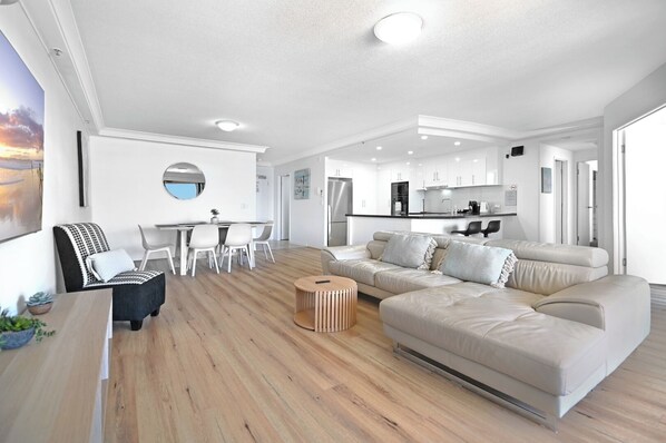 Ein modernes Wohnzimmer mit Sofa, Esstisch und Stühlen. Es gibt eine Küchenzeile mit Schränken und Geräten.