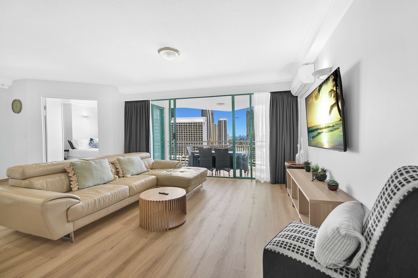 A modern living room with a beige sectional sofa, a round coffee table, and a view of a cityscape through large glass doors.