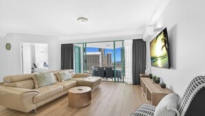 Ein modernes Wohnzimmer mit einem beigefarbenen Ecksofa, einem runden Couchtisch und Blick auf eine Stadtlandschaft durch große Glasschübe.