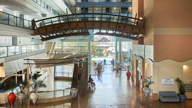 Ein großzügiger Innenbereich mit einer geschwungenen Glasbrücke, die zwei Ebenen verbindet, einem zentralen Atrium mit Sitzgelegenheiten und einem Schild, das einen Aufzugsbereich kennzeichnet.