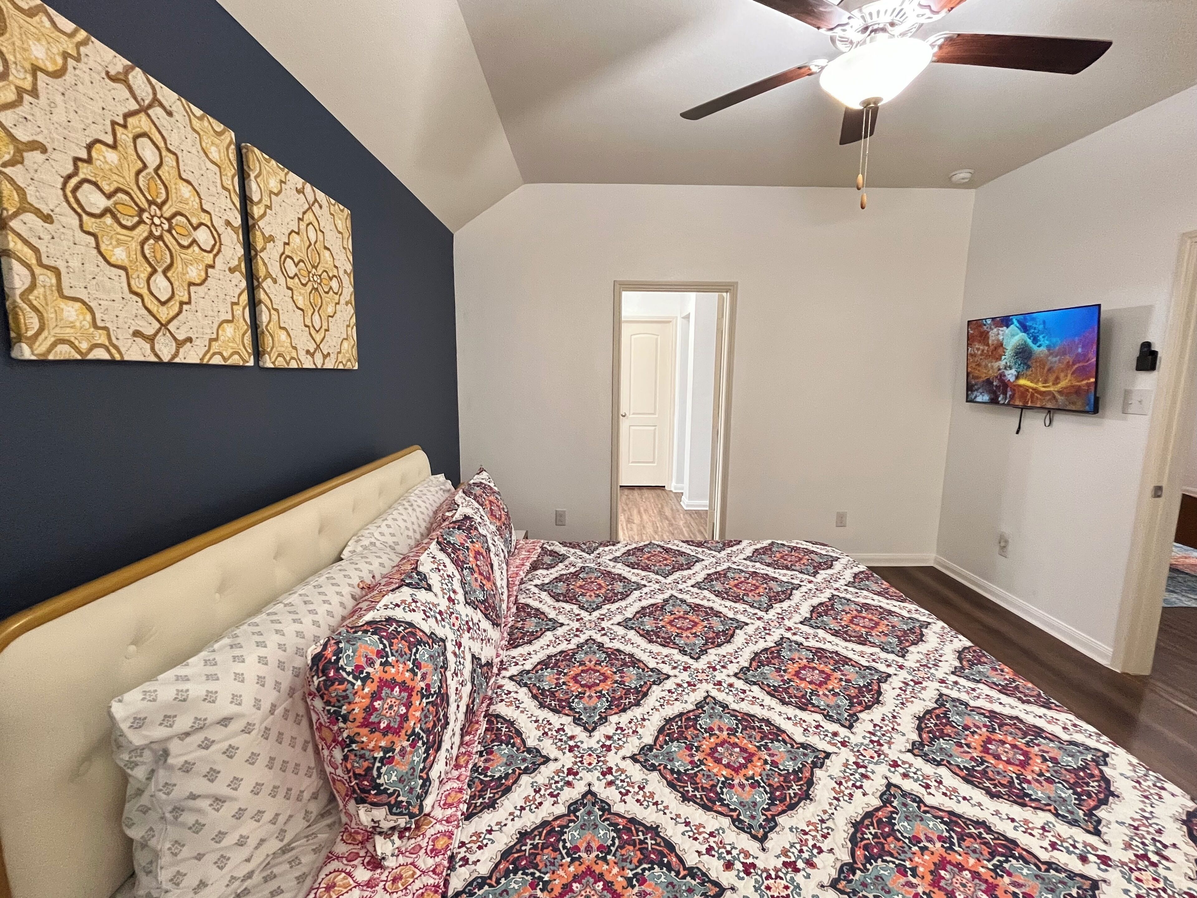 A bedroom with a large bed, a ceiling fan, a TV, and a door leading to another room.